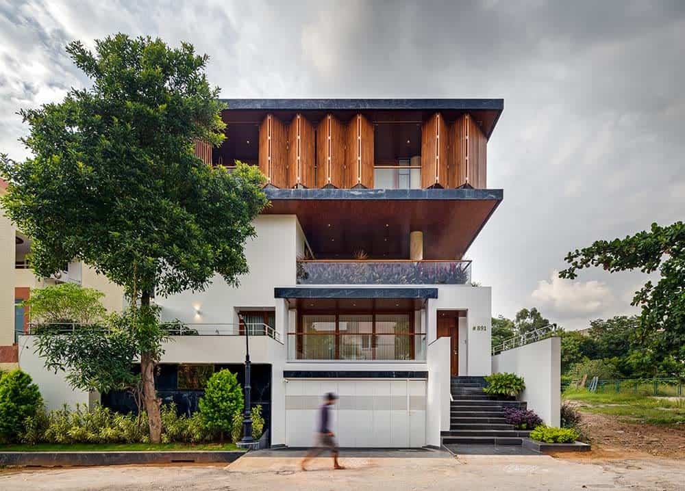 This is a front exterior view of the house with bright white exterior walls on the lower levels adorned by the tall tree and windows On the upper levels are balconies and wooden folding panels that open to glass walls
