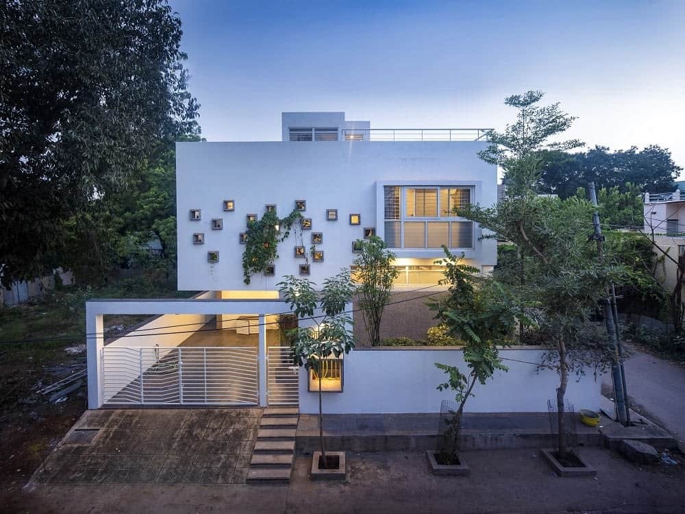 This is an aerial view of the front exterior of the house with white exterior walls complemented by the warm lighting as well as the creeping vines of the landscape that also has tall trees