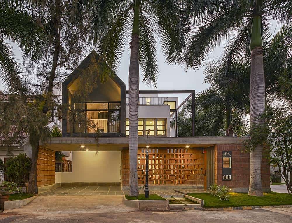 This is a front exterior view of the house with a alrge carport on one side topped with glass walls and large balconies These are then complemented by the patterned red brick walls and tropical trees